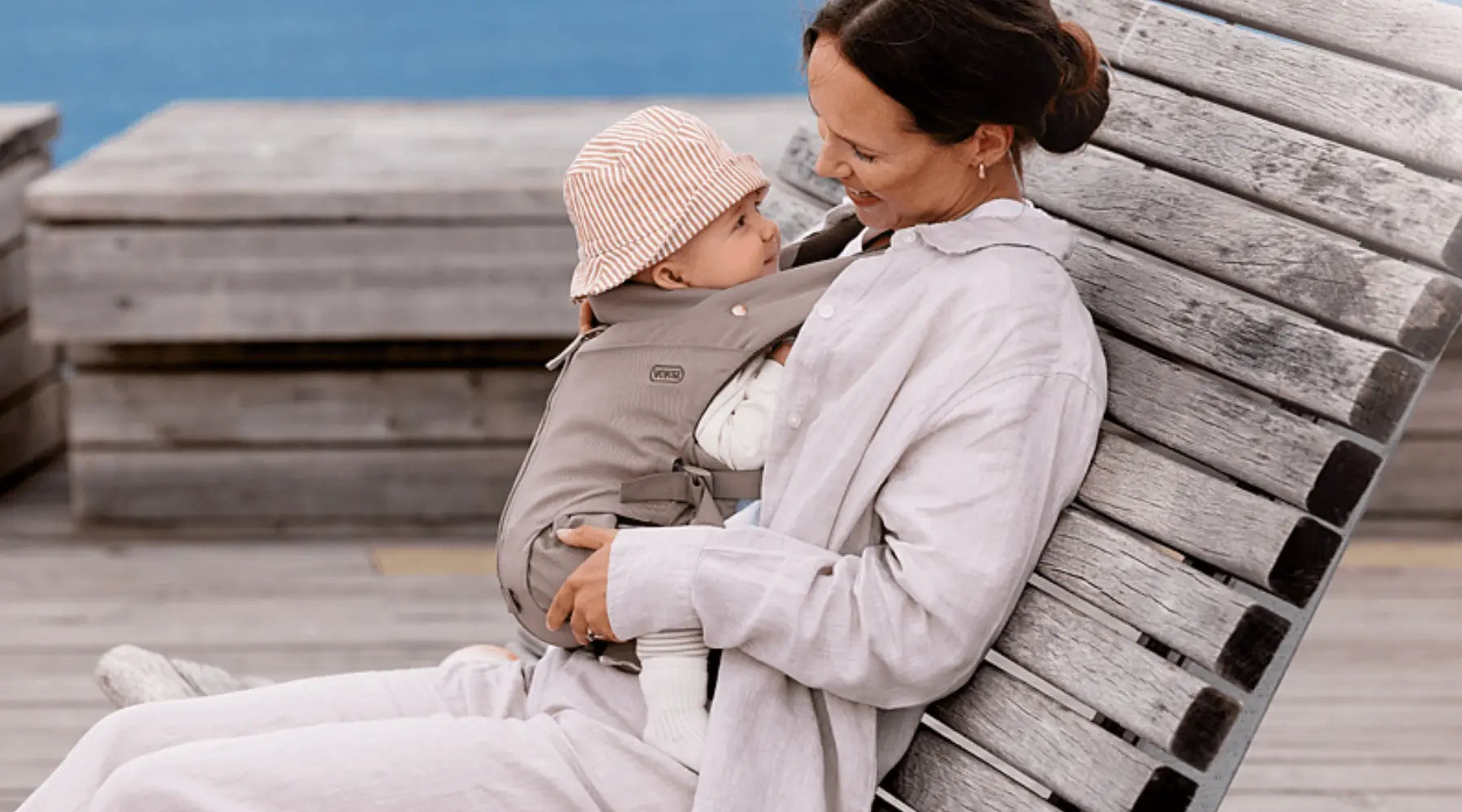 Eine sitzende Mutter lächelt ihr Baby an, das sie in der Voksi Babytrage Huddle Clay trägt, während sie sich auf einer Holzliege im Freien entspannt.
