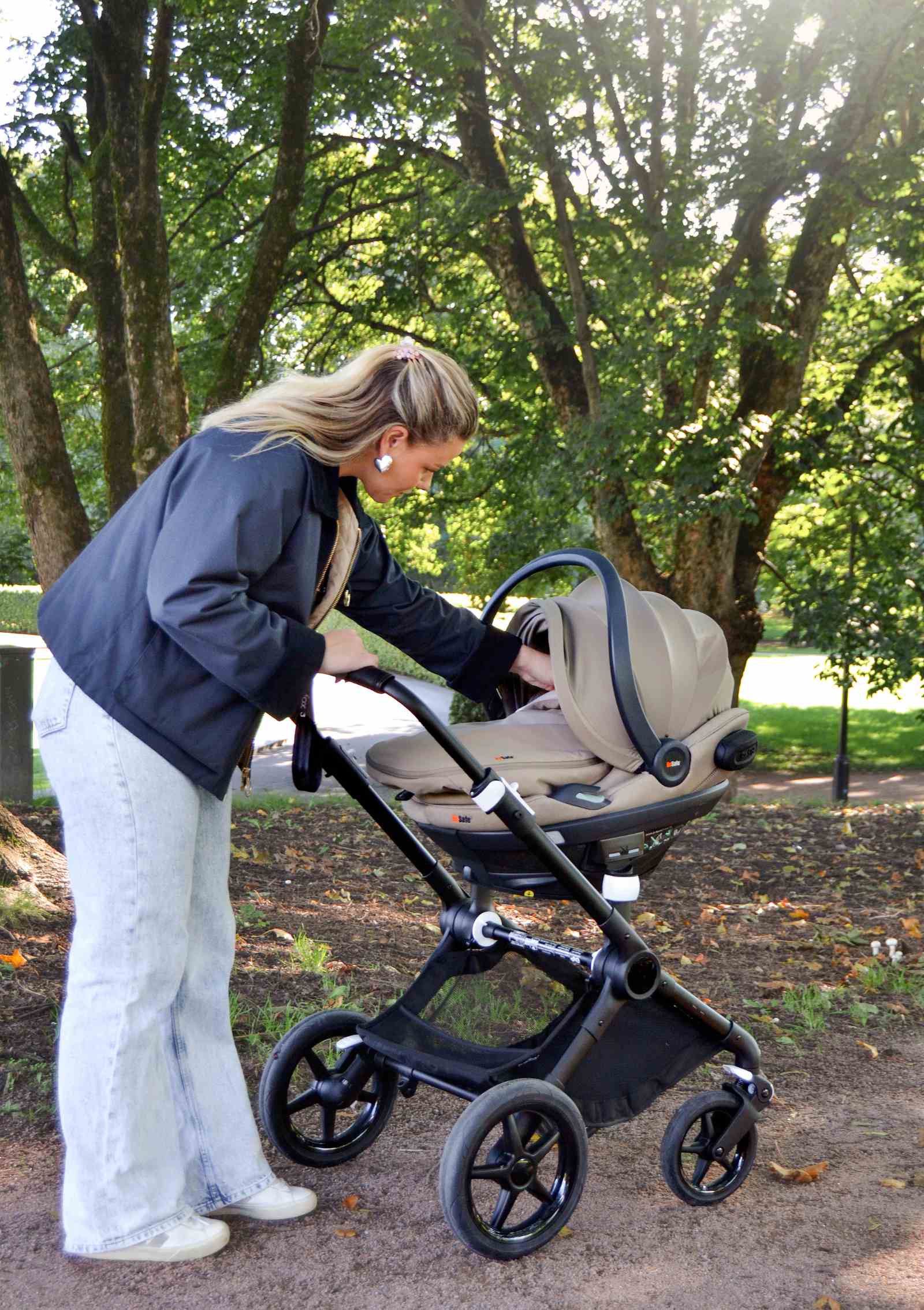 Frau schiebt am Kinderwagen befestigte BeSafe Babyschale Go Beyond, ausgestattet mit Wind & Weather Cover Dark Sand