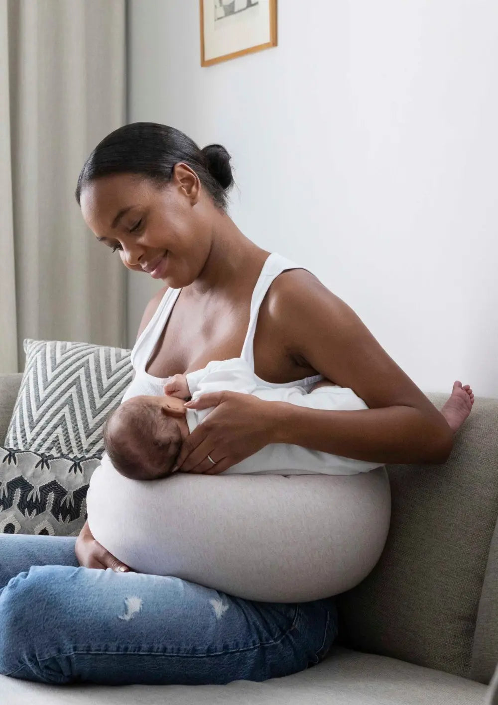 Lächelnde Mutter sitzt auf einem Sofa und stillt ihr Neugeborenes mit dem bbhugme Stillkissen Pink Mélange als Unterstützung.