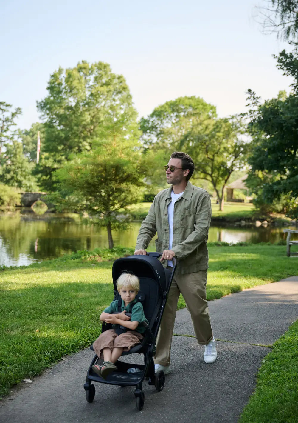 Ein Mann schiebt einen Nuna VIAA cabn Buggy mit einem Kleinkind darin auf einem asphaltierten Weg durch einen Park. Der Mann trägt eine grüne Jacke und Sonnenbrille. Im Hintergrund sind ein Teich und Bäume zu sehen. Das Bild zeigt den Buggy in voller Funktion und Einsatzbereitschaft im Alltag.