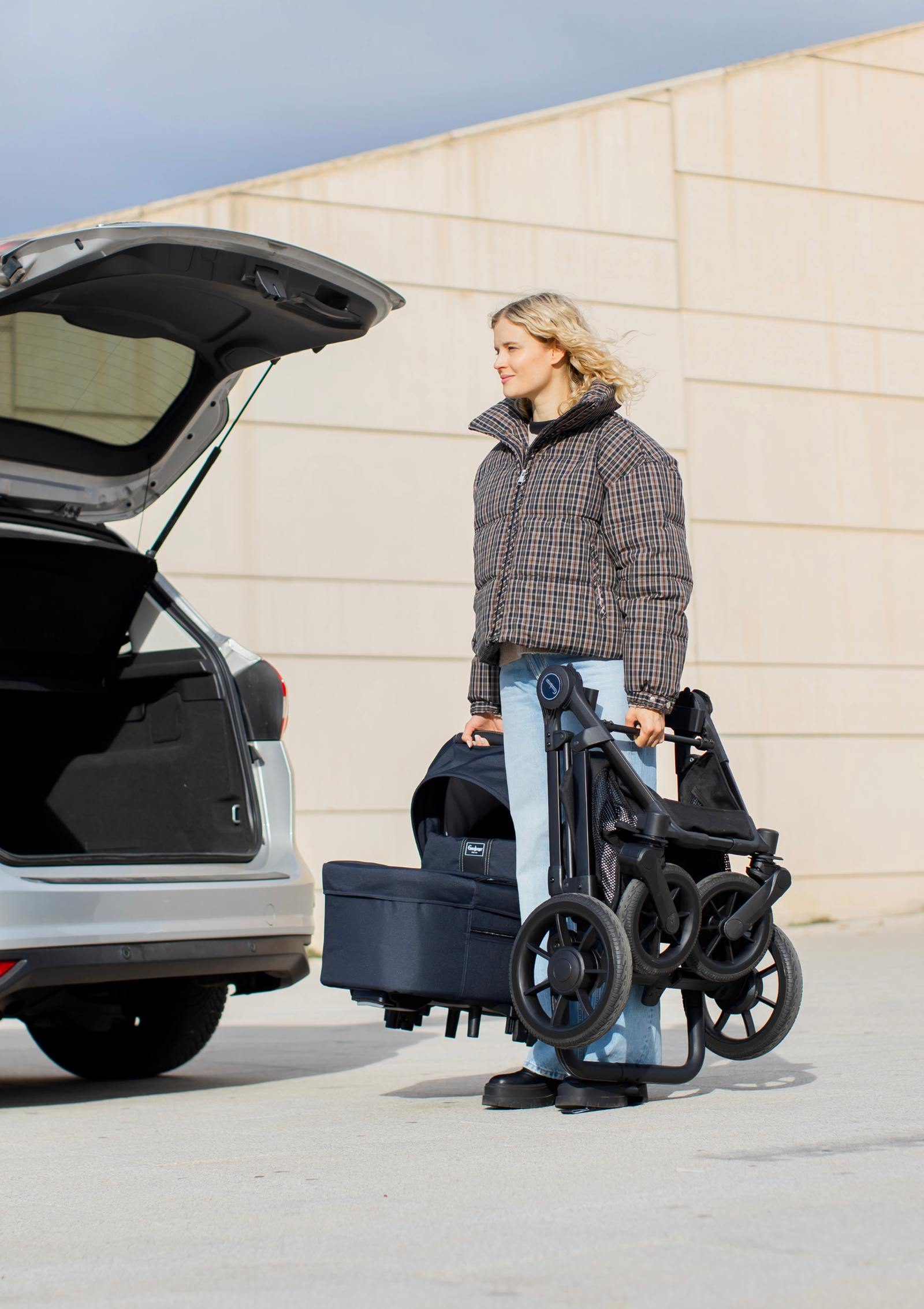 Frau verstaut zusammengeklappten Emmaljunga Kinderwagen 'SUPER SONIC' Urban Dark Navy im Kofferraum