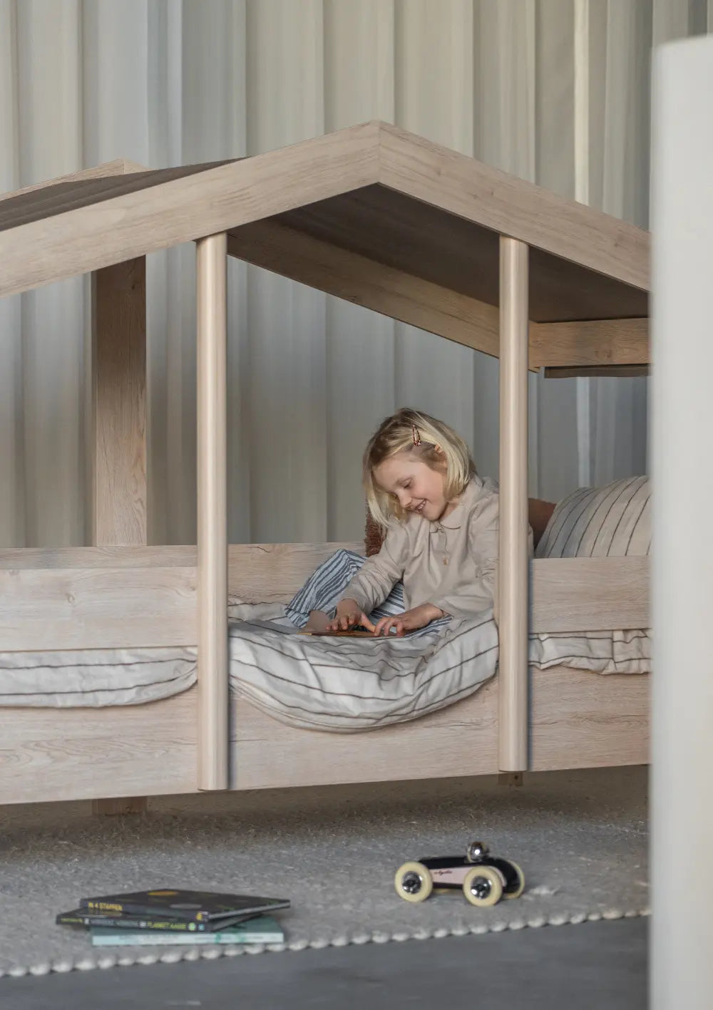 Ein lächelndes blondes Kind sitzt gemütlich im Quax Hausbett Cottage Natural Ash (200 x 90 cm) und blättert unter dem dunkelbraunen Stoffdach in einem Buch.