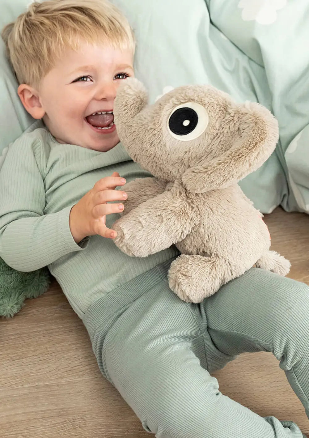 Ein glücklich lachendes Kleinkind liegt auf dem Rücken im Bett und hält den weichen, sandfarbenen Elefanten Done by Deer Kuschelfreund Elphee Sand mit seinen charakteristischen großen Kulleraugen im Arm