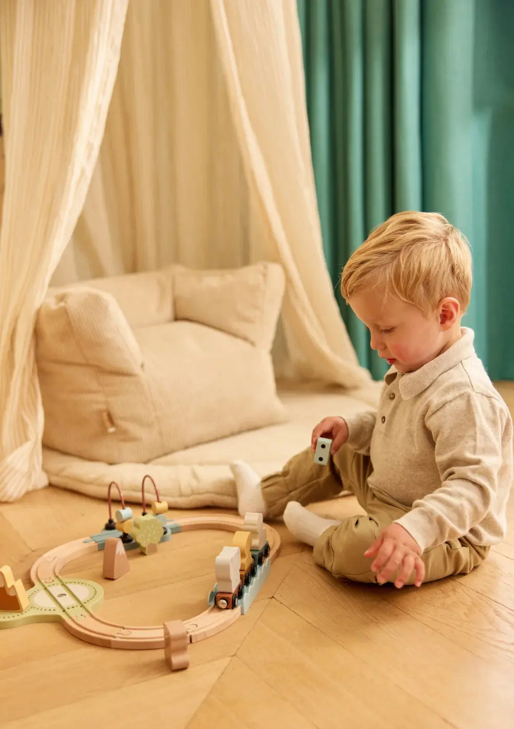 Ein Kleinkind sitzt auf dem Holzboden im Kinderzimmer und spielt zufrieden mit der kreisförmig aufgebauten Jollein Holz Eisenbahn Set Jollies.
