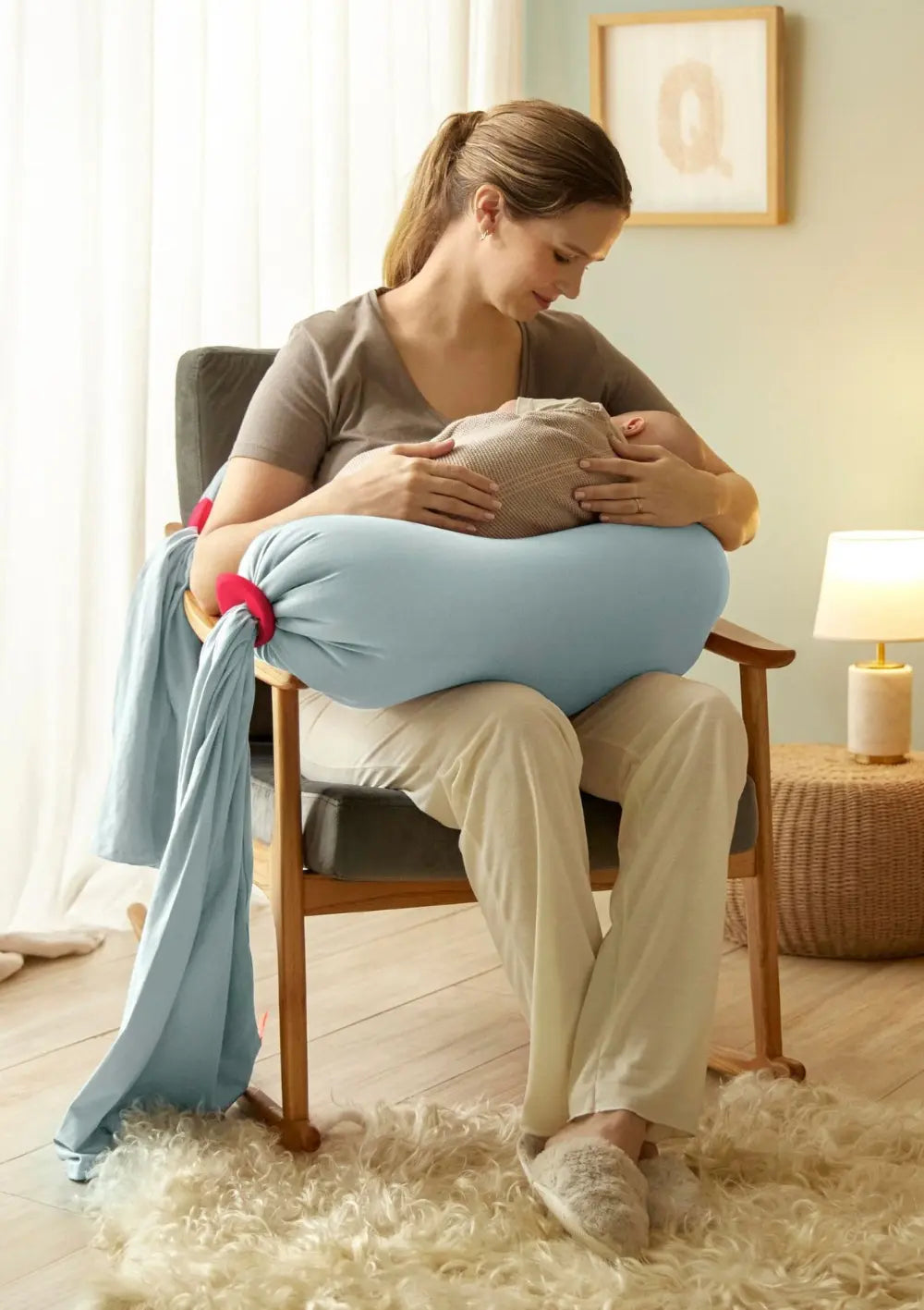 Eine Frau sitzt auf einem Stuhl mit ihrem Baby im Arm und wird gestützt vom bbhugme Schwangerschaftskissen Eucalyptus