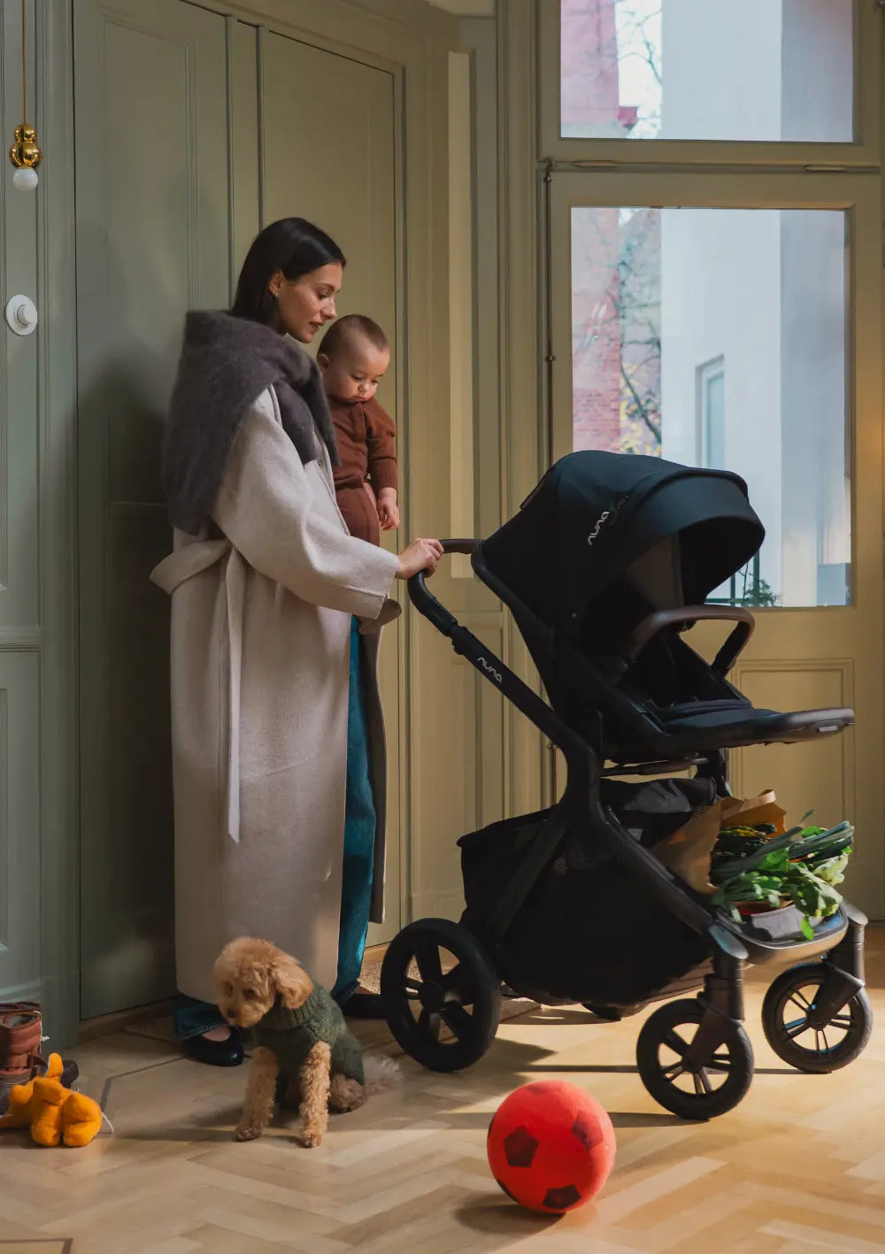 Frau mit Kind auf dem Arm schiebt Nuna Sportwagen 'DEMI icon' Caviar im Wohnungsflur, Einkäufe im extra großen Staukorb und Hund im Vordergrund
