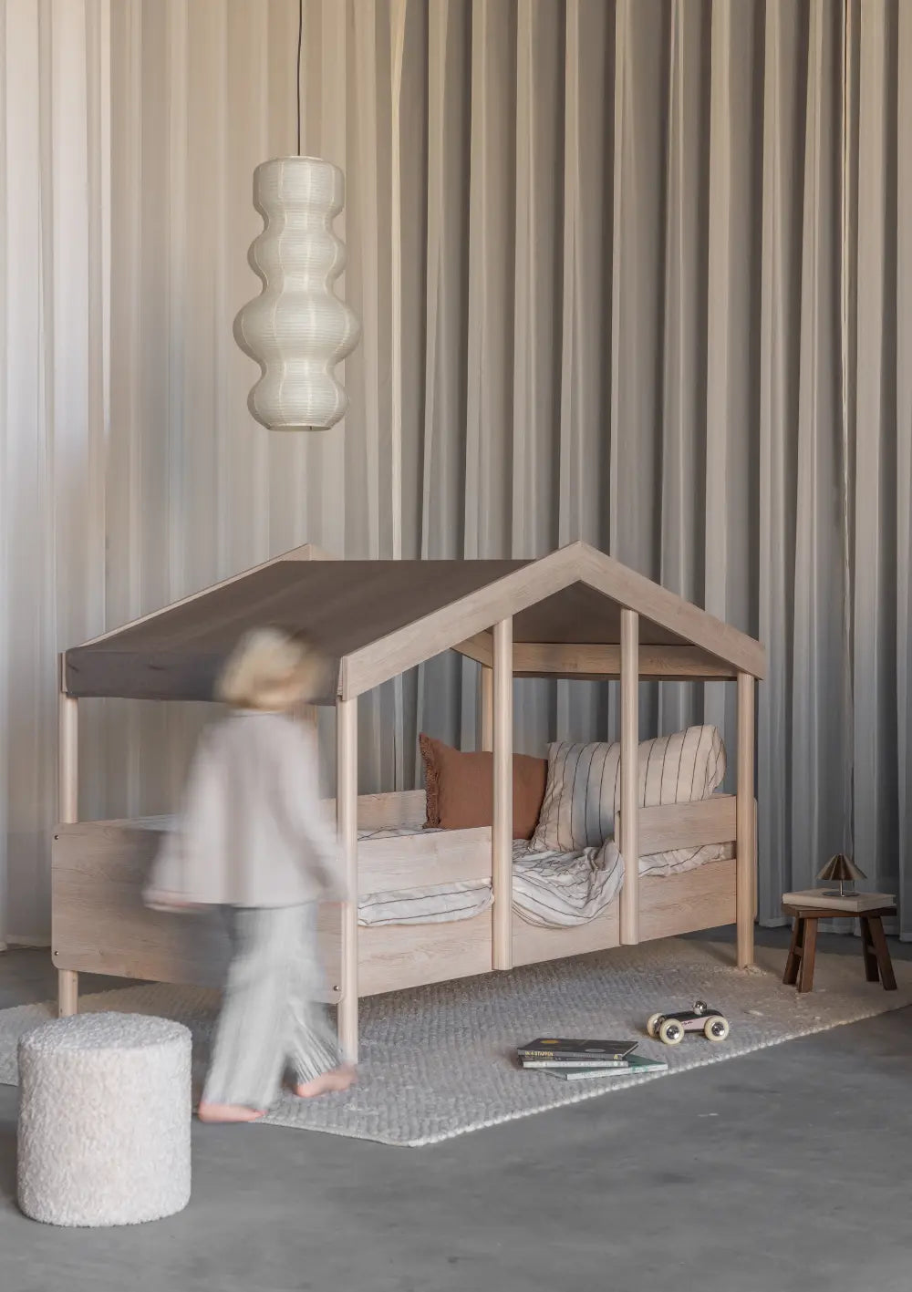 n Kind läuft an einem hellen Teppich im Kinderzimmer vorbei, auf dem das Quax Hausbett Cottage Natural Ash (200 x 90 cm) aus hellem Holz mit einem dunkelbraunen Stoff-Dachhimmel steht.
