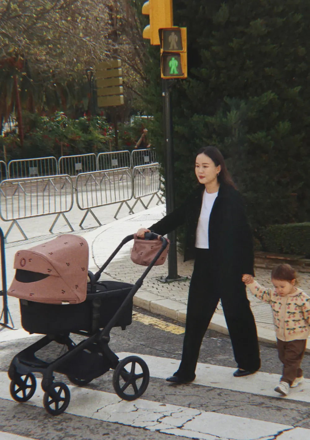 Eine Frau spaziert mit ihrem Kind über den Zebrastreifen; der Kombikinderwagen ist mit dem gemusterten Bugaboo Sonnendach Fox 5 Renew x Konges Sløjd Dusty Pink Cherry und der passenden Lenkertasche ausgestattet.