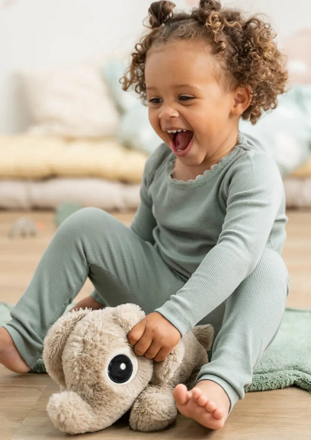 Ein fröhliches Kind mit Locken sitzt auf einem grünen Spielteppich und greift spielerisch nach dem plüschigen, beigen Elefanten Done by Deer Kuschelfreund Elphee Sand.