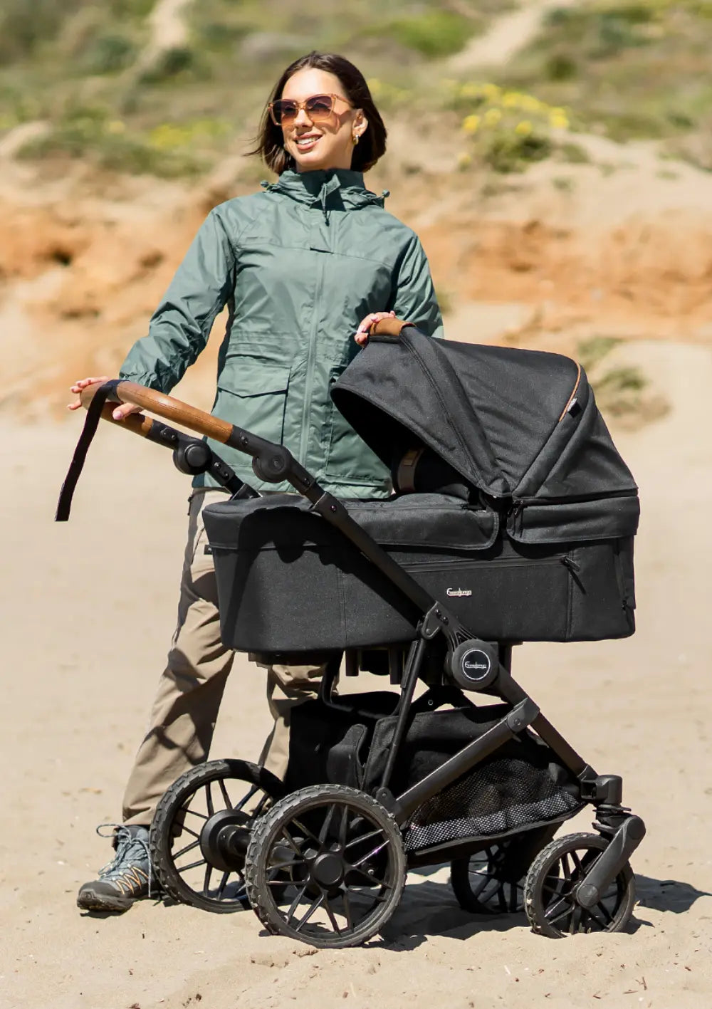 Eine Frau in einer grünen Outdoor-Jacke schiebt den schwarzen Kombikinderwagen mit Babywanne Emmaljunga 'SUPER SONIC' Urban Black über einen sandigen Weg im Freien.