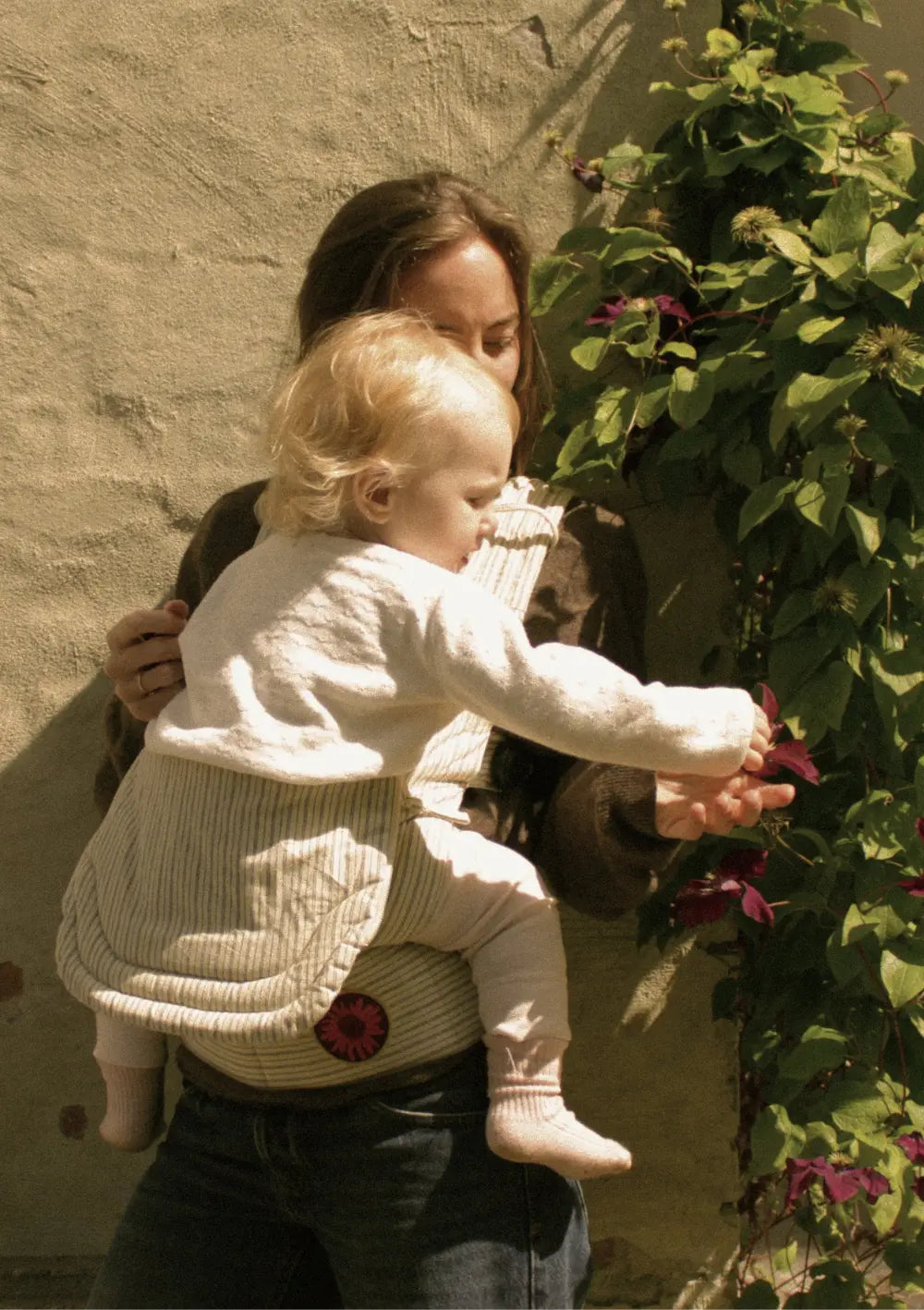 Eine Mutter trägt ihr Kleinkind auf der Hüfte in der beige gestreiften Moova Babytrage Balance Fine Lines, während das Kind im Freien neugierig nach einer lila Blüte an einer Pflanze greift.