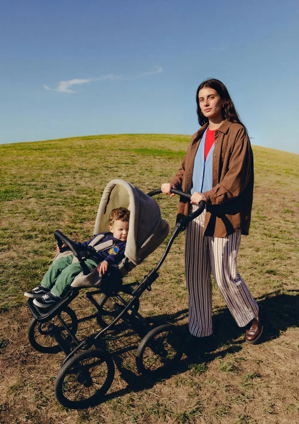 Mutter schiebt den Angelcab Kinderwagen 'C3-Series' Upminster mit Kleinkind im Sportsitz über eine sonnige Wiese