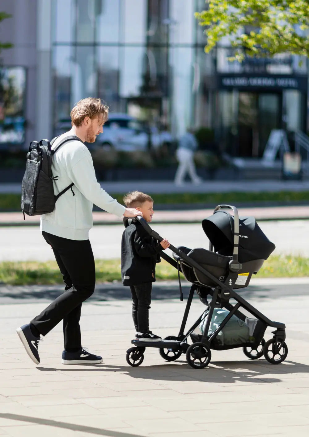Vater schiebt Kinderwagen mit aufgesetzter Babyschale, ausgestattet mit dem Thule Mitfahrbrett 'Rider Board', auf dem ein Junge steht