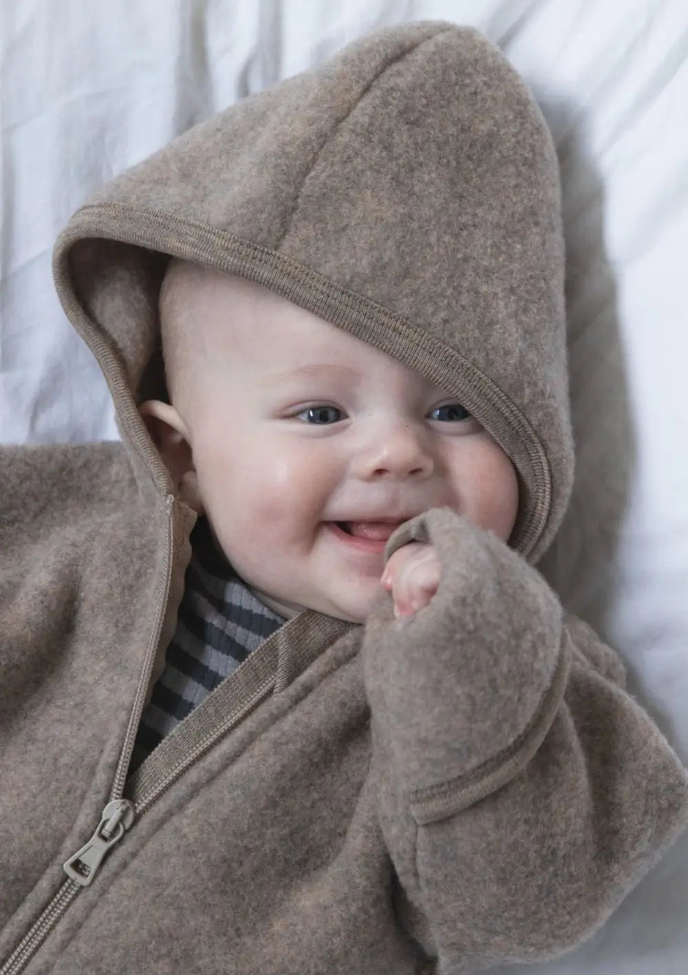 Lächelndes Baby liegt mit dem Engel Wollfleece Zip Overall mit Kapuze auf dem Bett