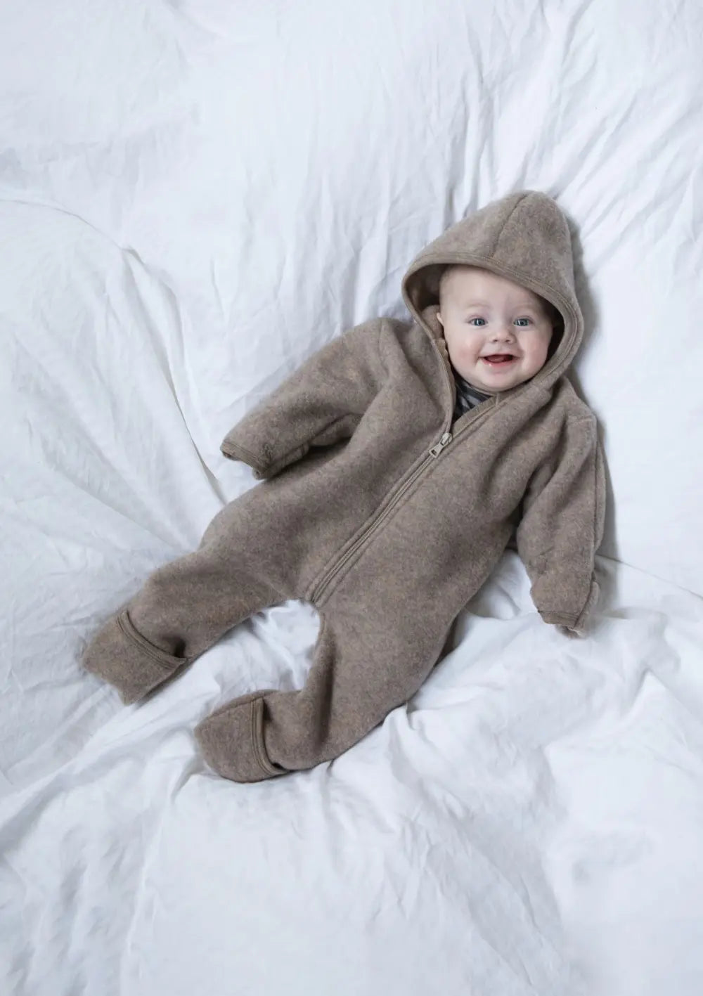 Lächelndes Baby liegt mit dem Engel Wollfleece Zip Overall mit Kapuze auf dem Bett