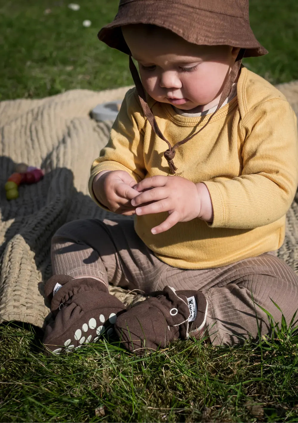 Baby sitzt auf einer Decke im Park und trägt die Kings & Rebels Booties Gamuza Chocolate